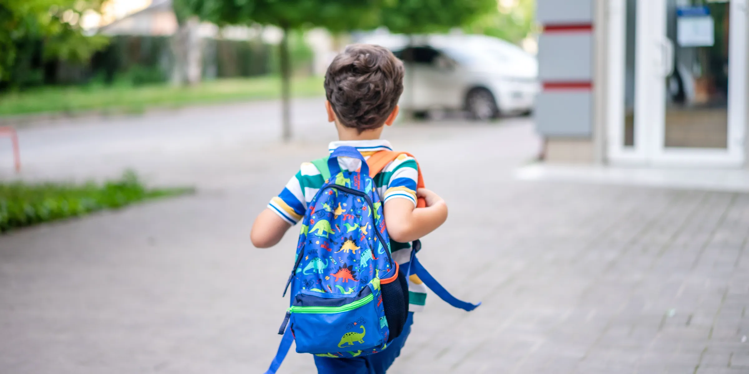 Grundschulkind mit buntem Rucksack geht über einen gepflasterten Gehweg auf einen Gebäudeeingang zu