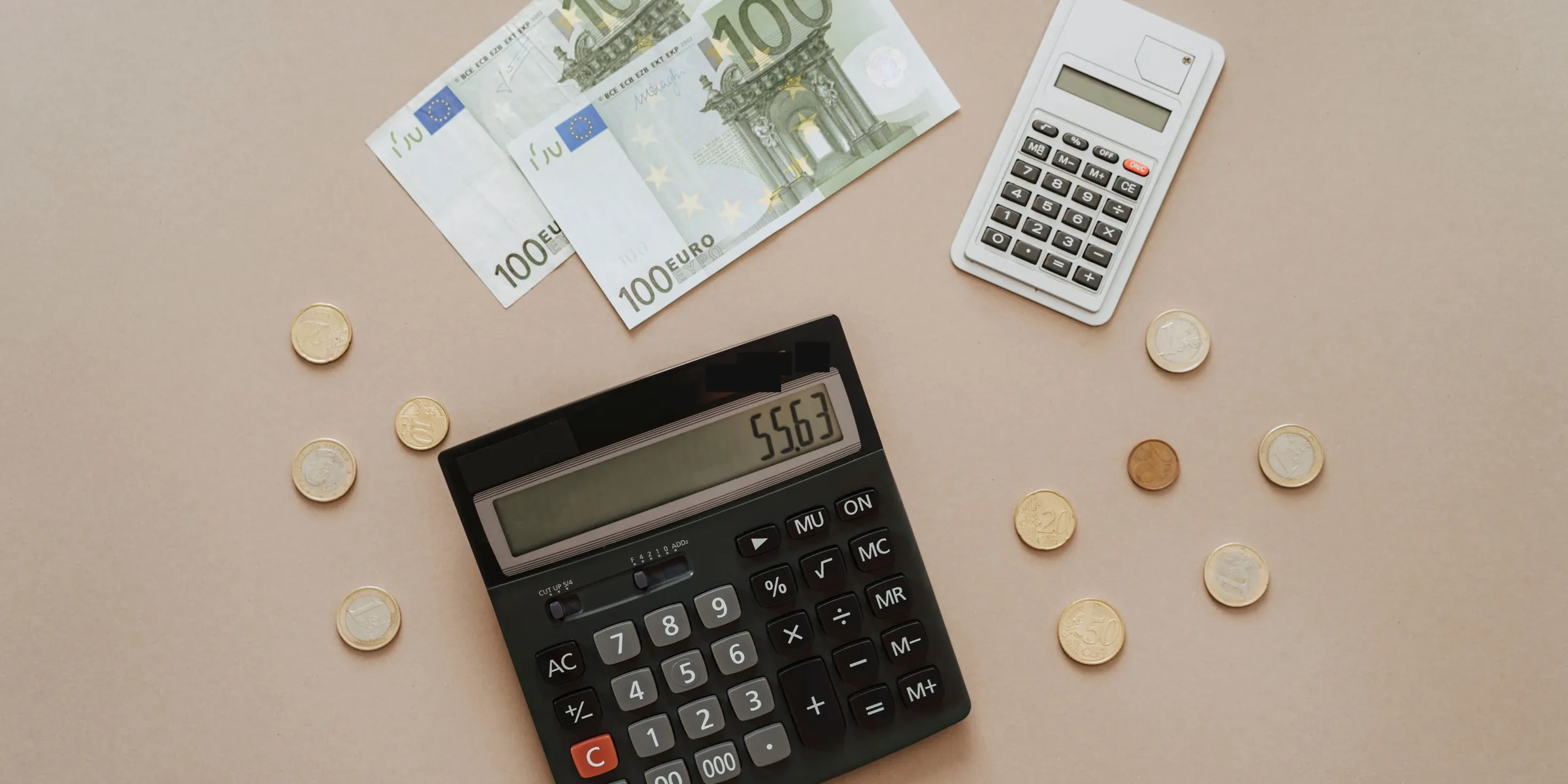 Zwei 100-Euro-Scheine, Euromünzen und zwei Taschenrechner liegen auf einer hellen Fläche