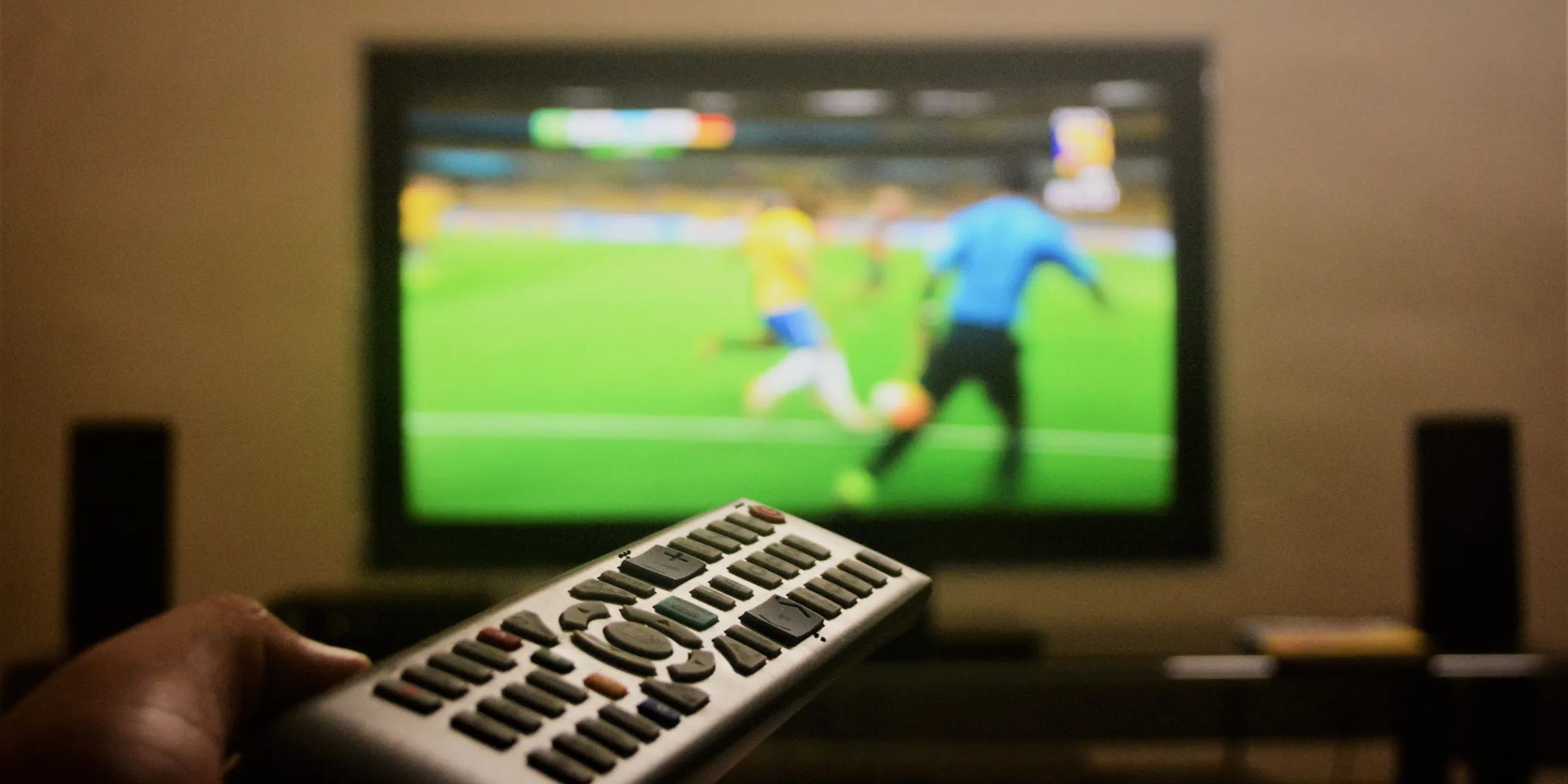 Eine Hand hält eine Fernbedienung vor einen Fernseher, auf dem gerade ein Fußballspiel übertragen wird