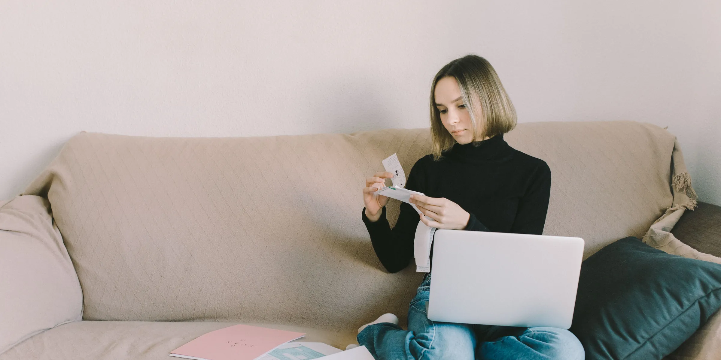 Frau sitzt mit Laptop auf dem Sofa und prüft Unterlagen, neben ihr liegen Formulare und Hefte ausgebreitet