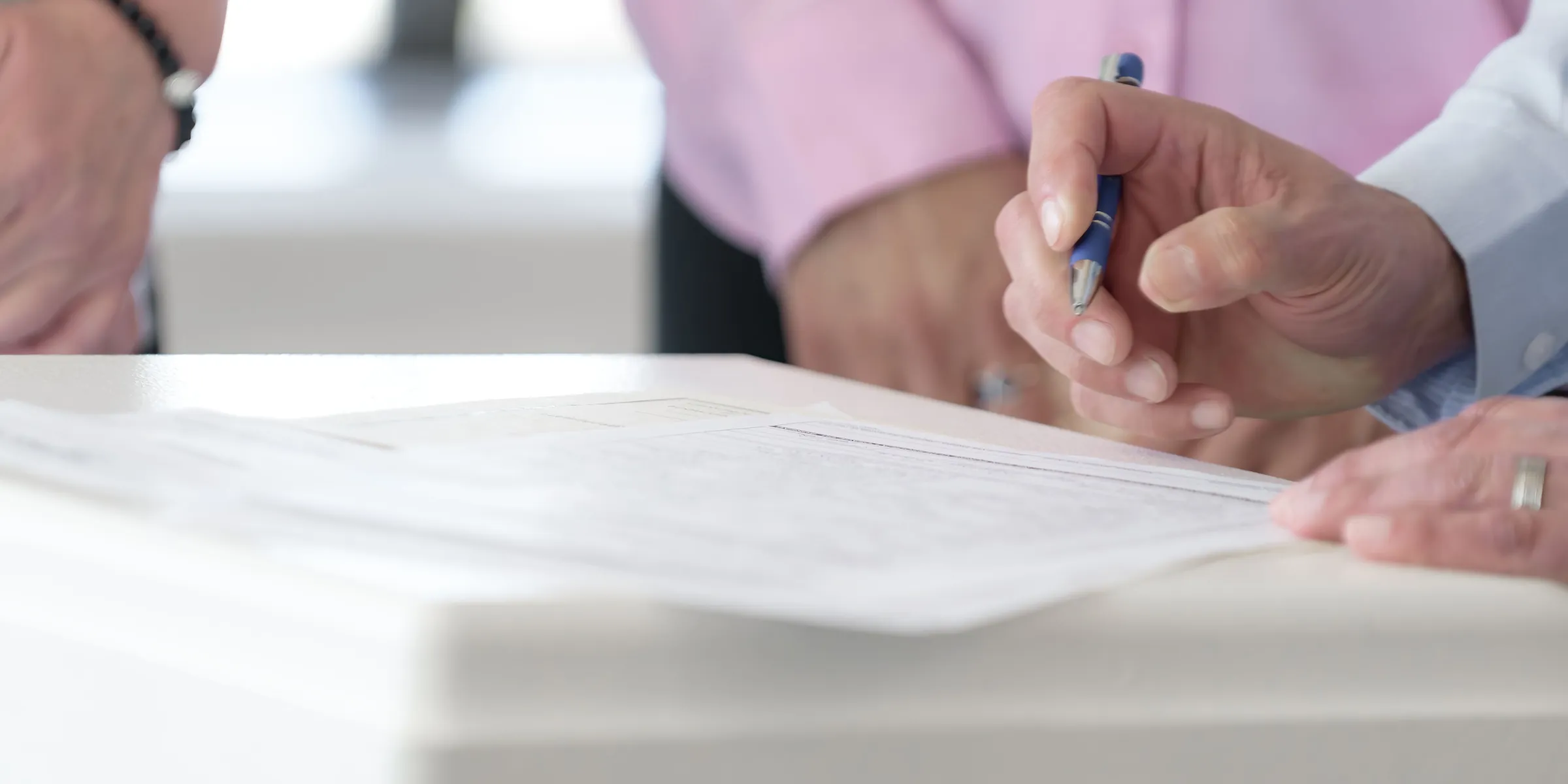 Hand mit Kugelschreiber setzt zur Unterschrift auf einem Formular an, eine zweite Person steht dabei am Tisch