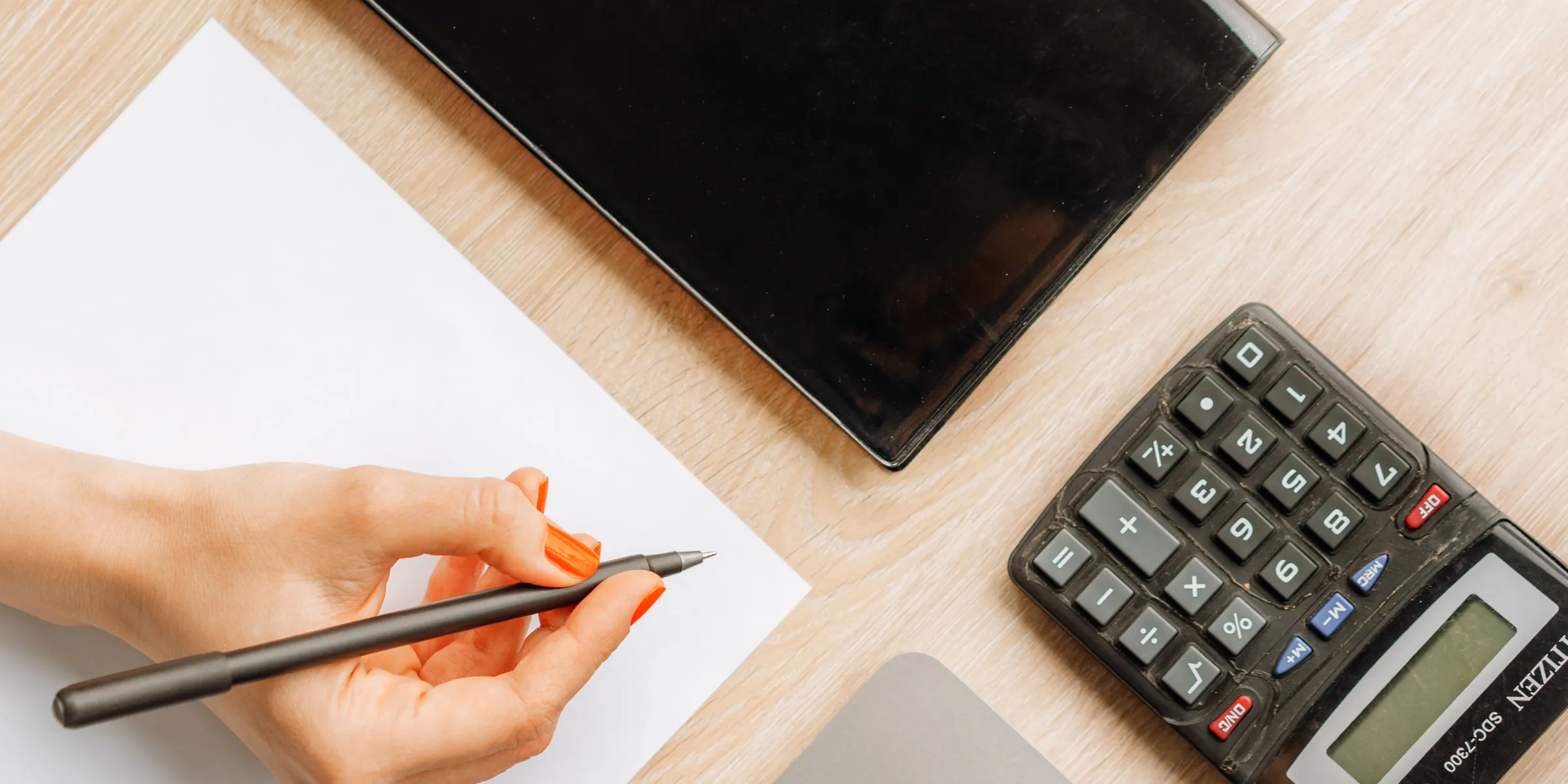 Hand mit Stift über einem Blatt Papier, daneben ein Taschenrechner und ein Tablet auf einem hellen Holzschreibtisch