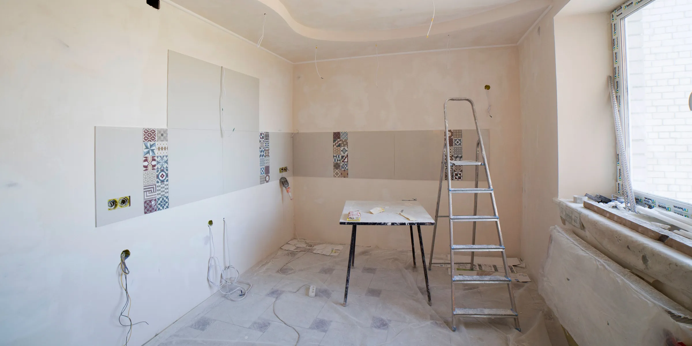 Leerer Raum in der Renovierung mit abgedecktem Boden, Trittleiter, Arbeitstisch und angefangenen Fliesen an der Wand